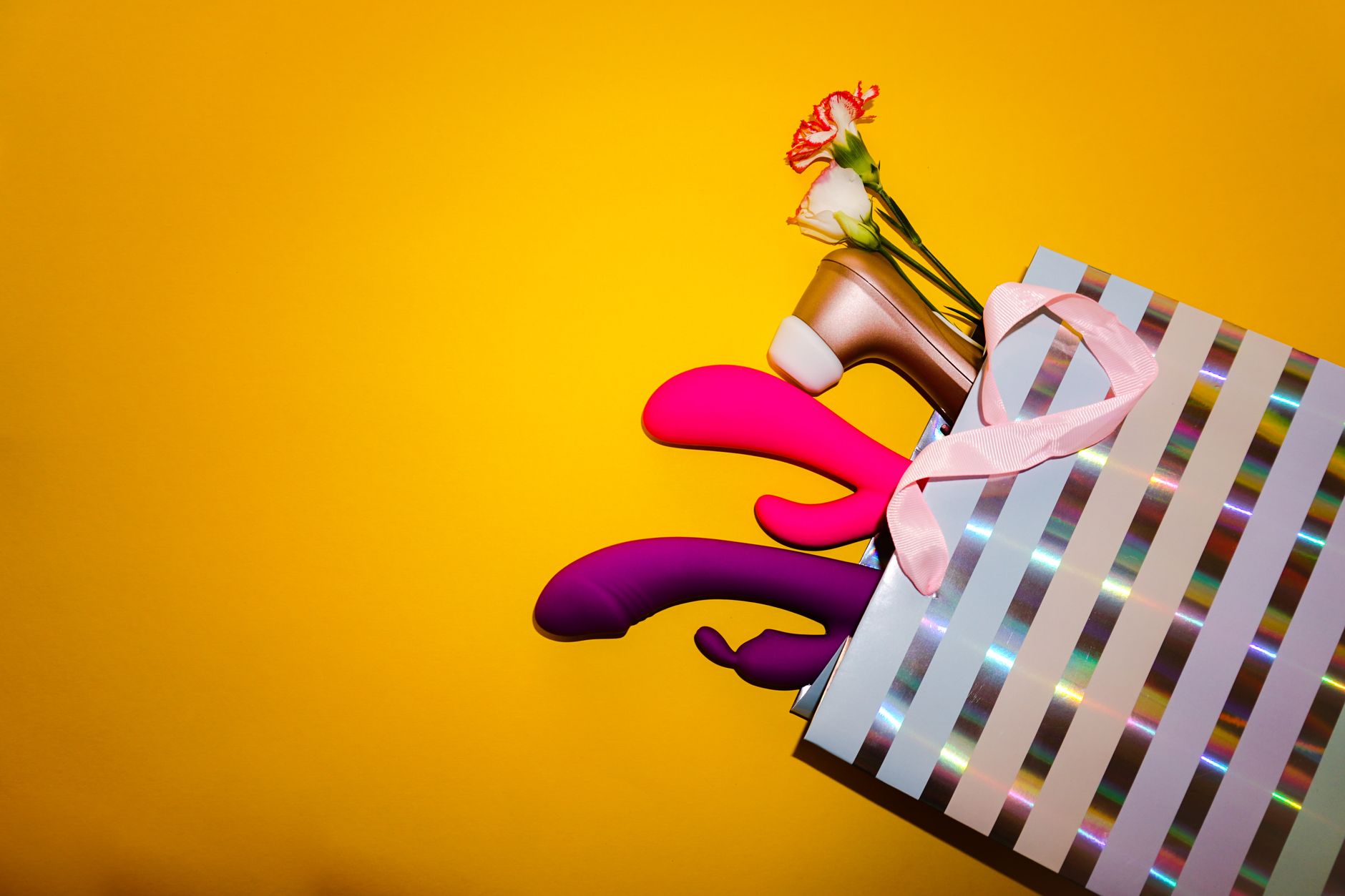 Colorful clitoral vibrators displayed with flowers in a gift bag against a bright yellow background